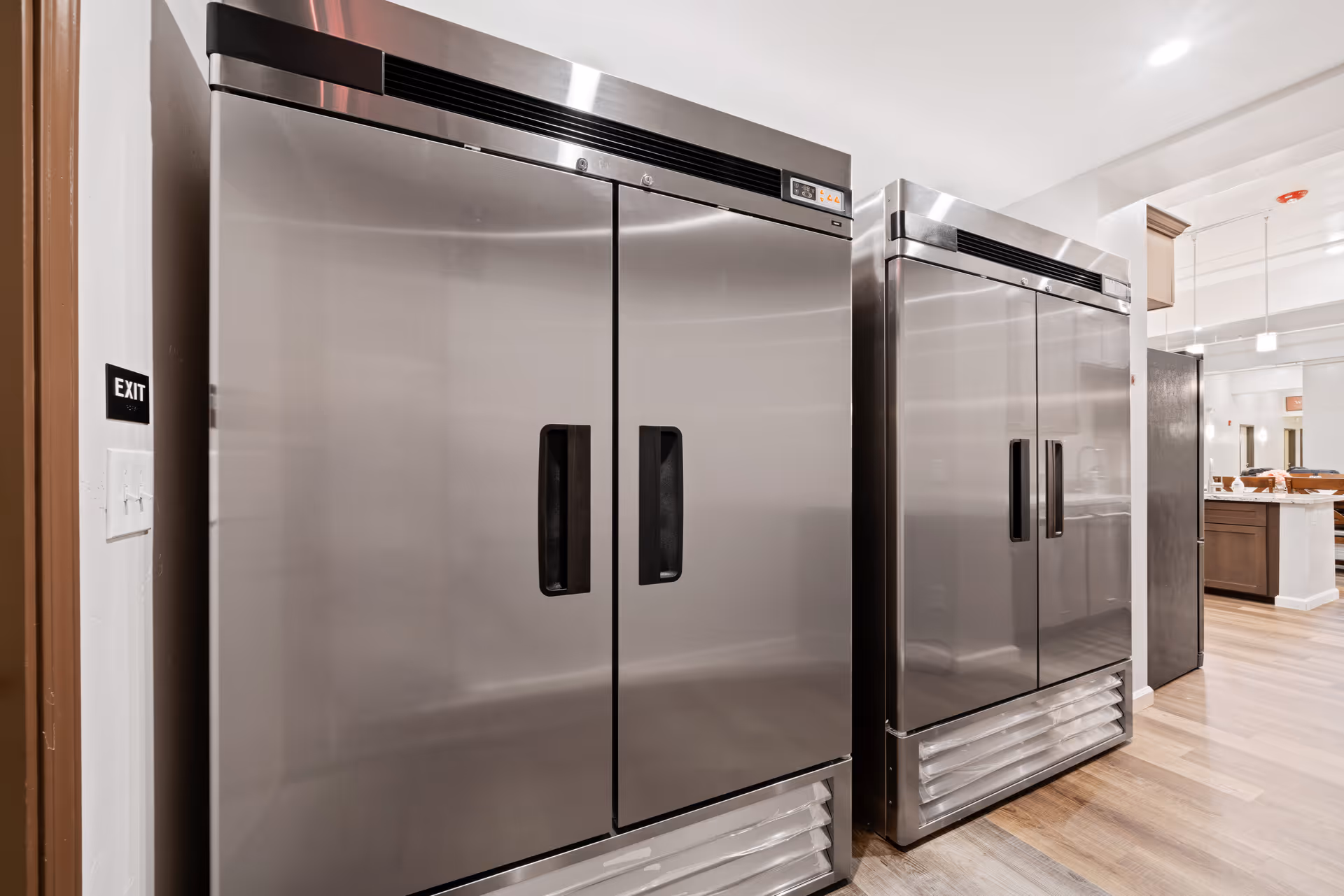 Two large stainless steel commercial refrigerators standing side by side in a kitchen area with wooden flooring and white walls. The kitchen area is well-lit with ceiling lights and has a modern design.