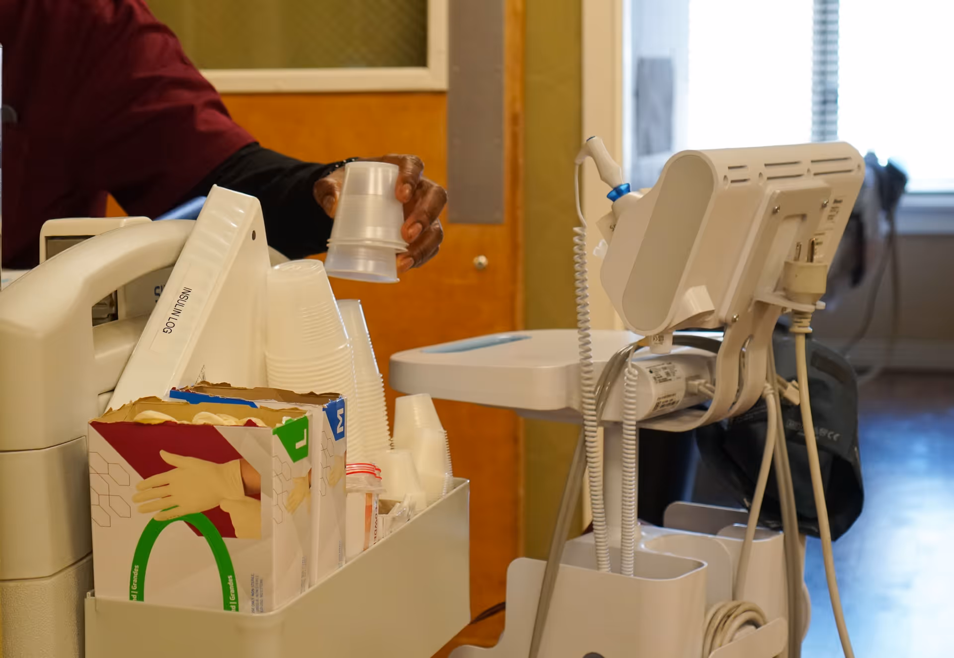 A close-up view of a medical cart with a blood pressure monitor and disposable plastic cups. A person wearing a maroon uniform is reaching for a cup. There is a box of disposable gloves labeled 'INSULIN LOG' on the cart. The background shows a door and a window with blinds.
