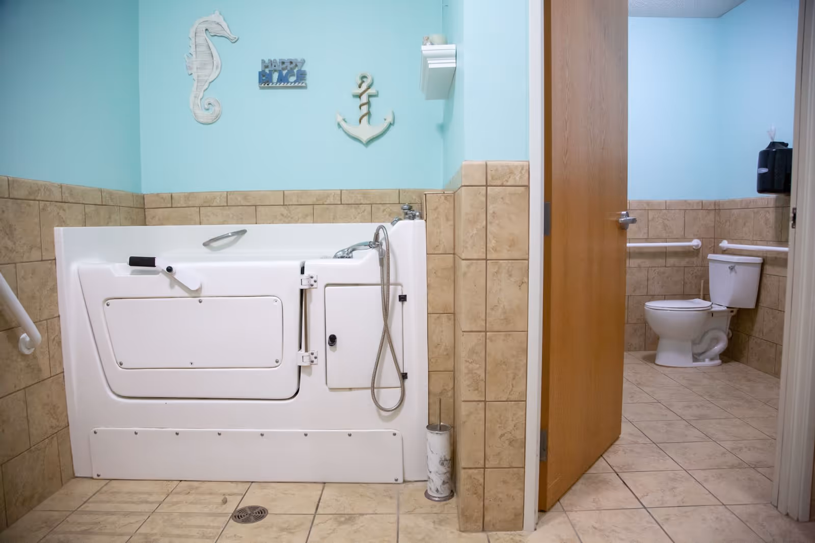 A bathroom with beige tiled walls and floor, featuring a white walk-in bathtub with a handheld showerhead on the left side. On the right, through an open wooden door, there is a toilet with grab bars on the walls. The walls are painted light blue and decorated with a seahorse, an anchor, and a 'Happy Place' sign.