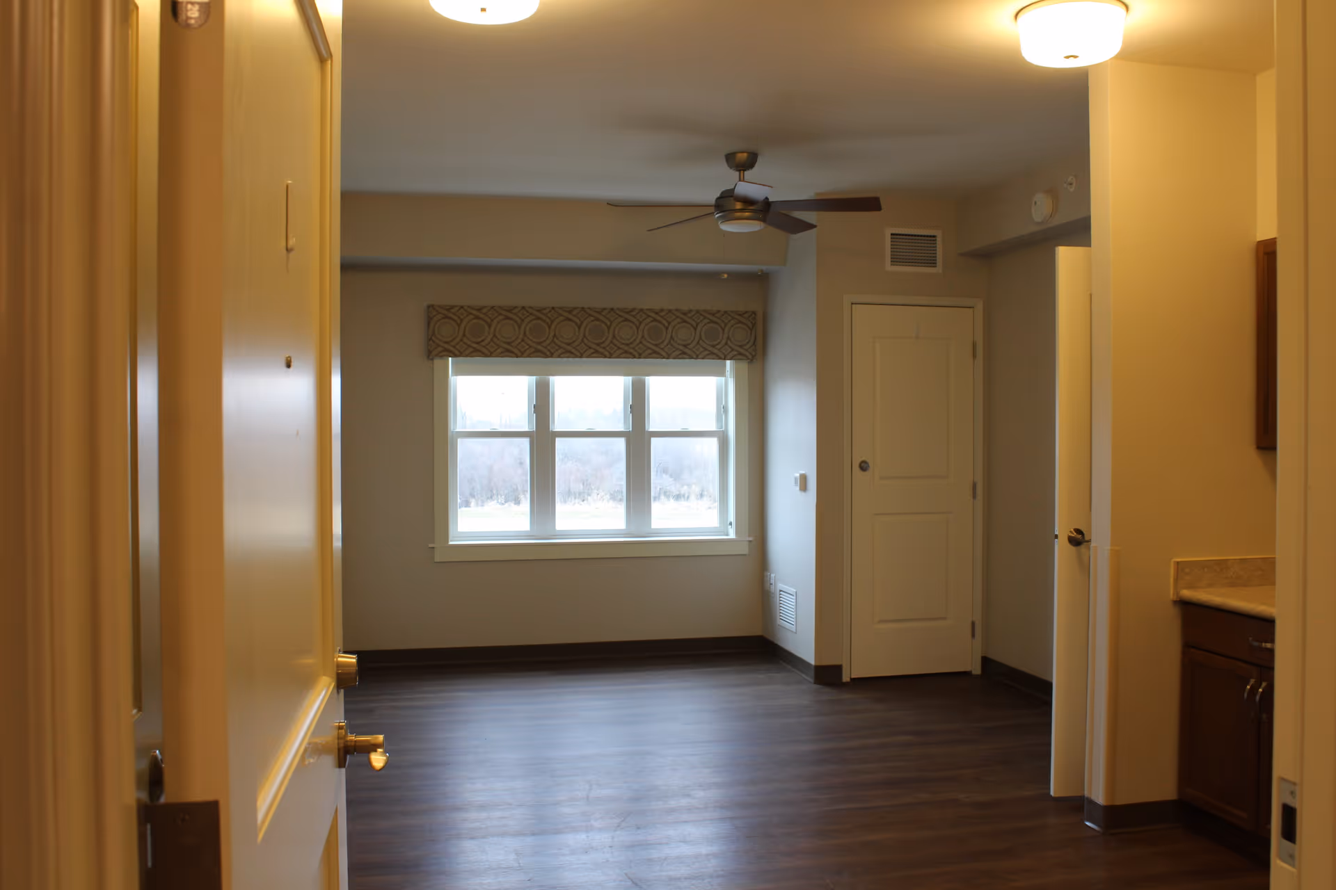 View into an empty room from an open doorway in a senior living facility. The room has wood flooring, a large window with a patterned valance, a ceiling fan, and neutral-colored walls. To the right, there is a partial view of a countertop and cabinets, and a closed door is visible in the back corner.