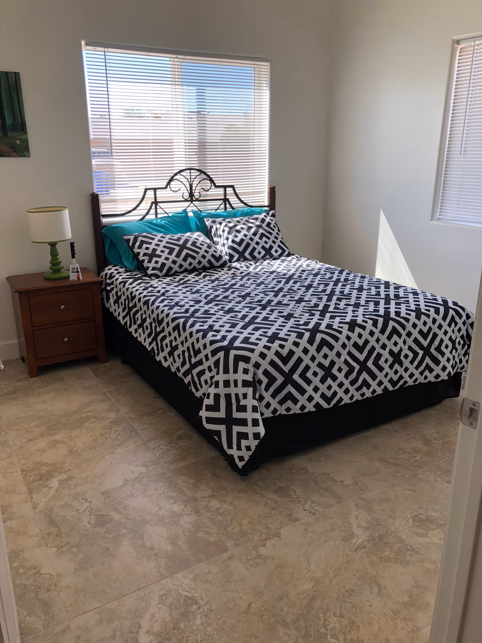 A bedroom with a bed featuring a black and white geometric patterned bedspread and two matching pillows, along with two teal pillows. The bed has a decorative metal headboard. Next to the bed is a wooden nightstand with two drawers, a green lamp, and a small bottle of hand sanitizer. The room has two windows with blinds, letting in natural light, and beige tiled flooring.