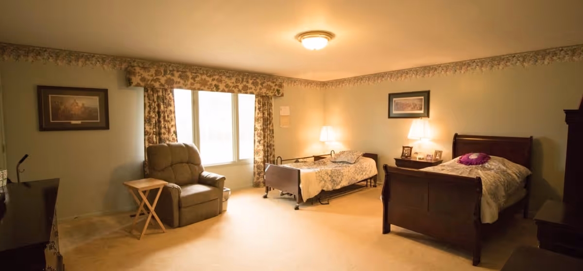 Spacious bedroom with two single beds, bedside tables and lamps, a recliner by the window, and warm overhead lighting.