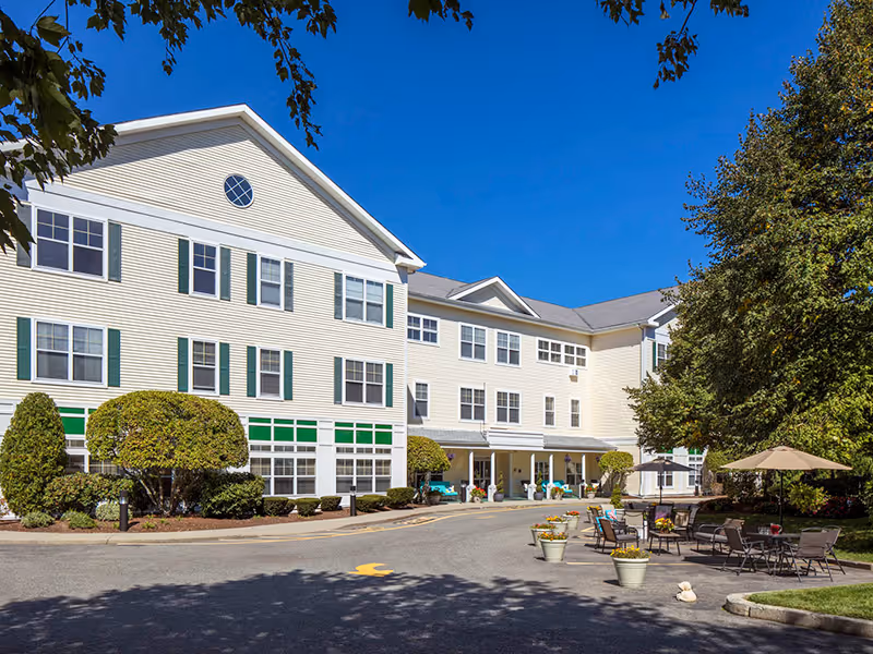 Three-story pale yellow senior living building with a covered entrance, landscaped shrubs, and outdoor seating with umbrellas.