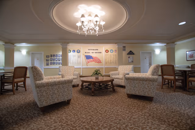 A cozy seating area in a senior living facility with patterned armchairs arranged around a round wooden coffee table. The room features a chandelier hanging from a decorative ceiling medallion. On the far wall, there is a 'Veterans Wall of Honor' display with an American flag and various plaques. The room has soft lighting and carpeted floors, with additional chairs and tables along the walls.