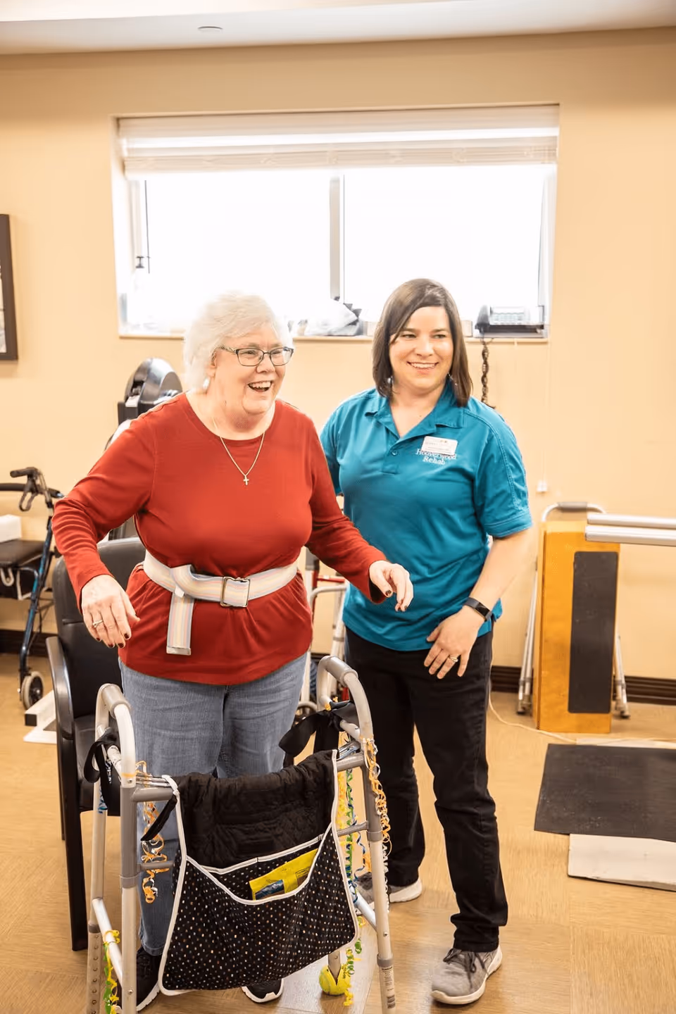 An elderly woman using a walker with a supportive belt around her waist is assisted by a smiling female caregiver wearing a teal shirt with a name tag. They are in a well-lit room with exercise equipment and a window in the background.