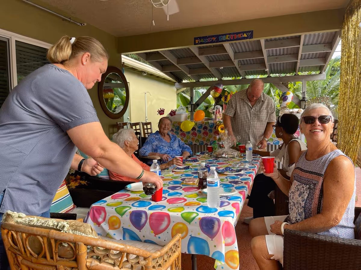 People, mostly older adults, sit around a balloon-themed table on a covered patio celebrating a birthday.