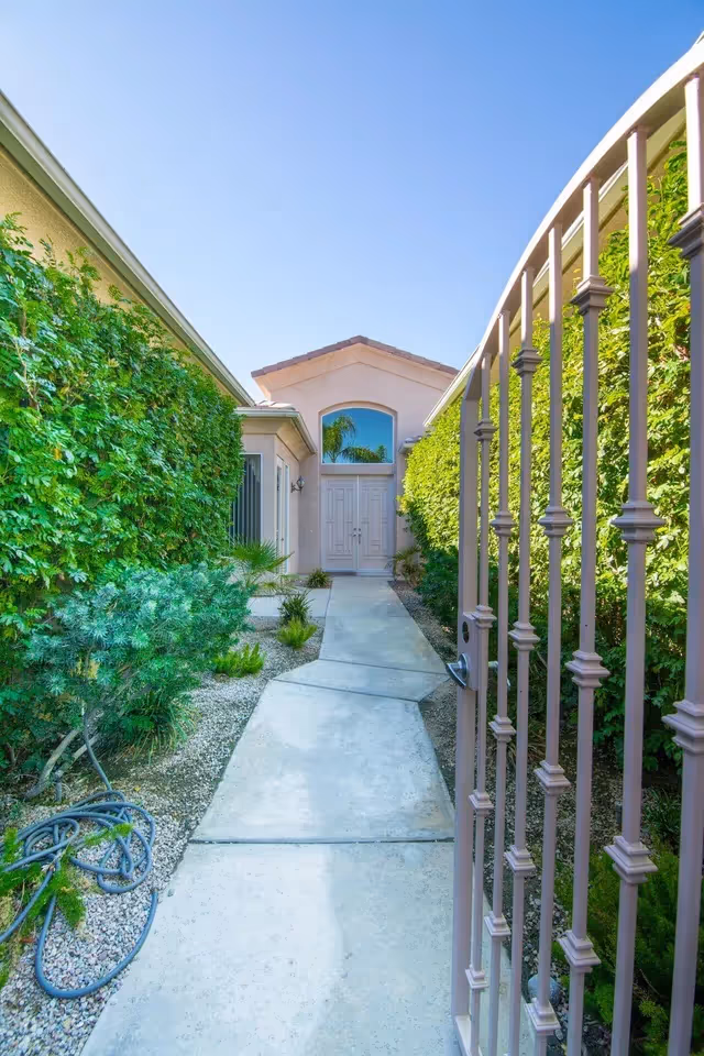 A concrete pathway leading to a front door of a building, flanked by green hedges and plants on both sides, with a metal gate partially open on the right side under a clear blue sky.