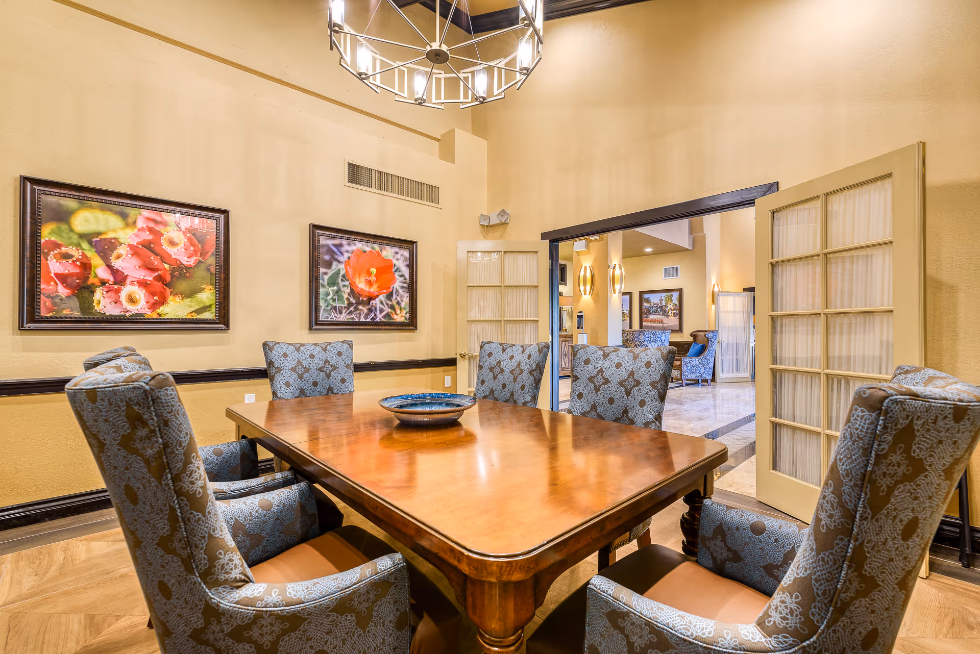 Bright dining room with a wooden table surrounded by upholstered chairs, floral artwork on the walls, and French doors opening to a lounge area.