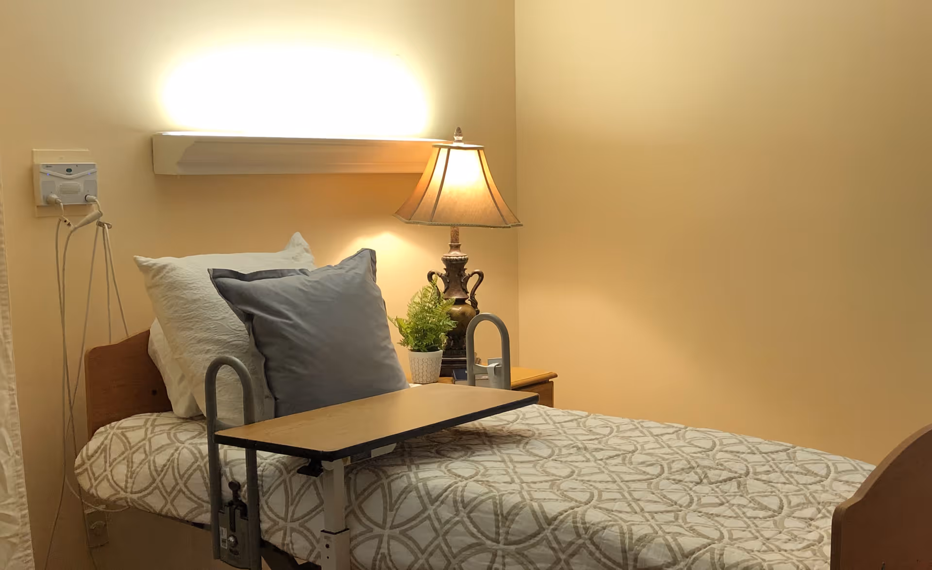 A neatly made single hospital-style bed with pillows, an overbed table, bedside lamp, and small plant in a softly lit room.