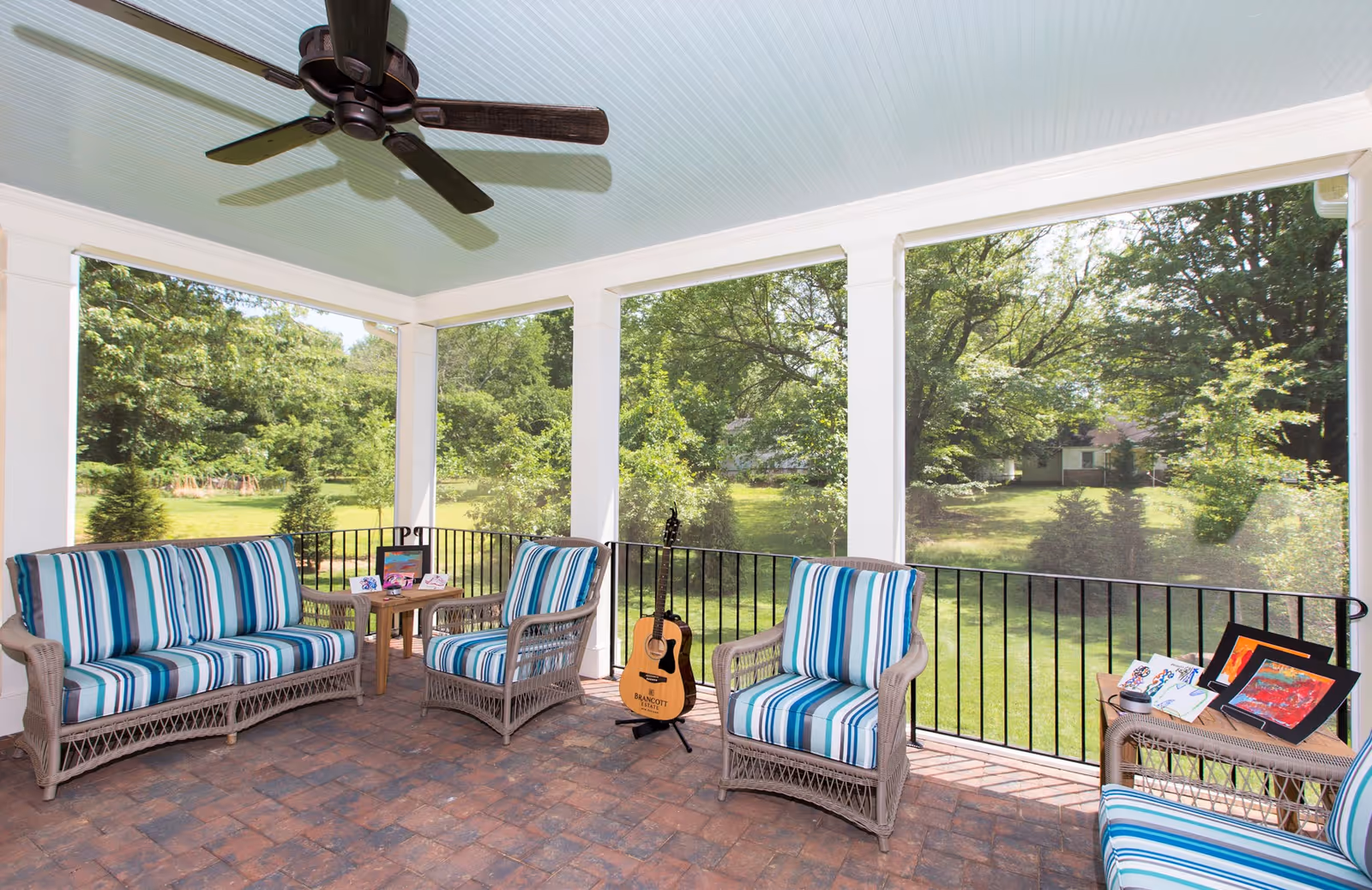 A screened-in porch with wicker furniture featuring blue and white striped cushions. There is a ceiling fan above and a guitar resting on a stand near the railing. The porch overlooks a green yard with trees and a house in the background.