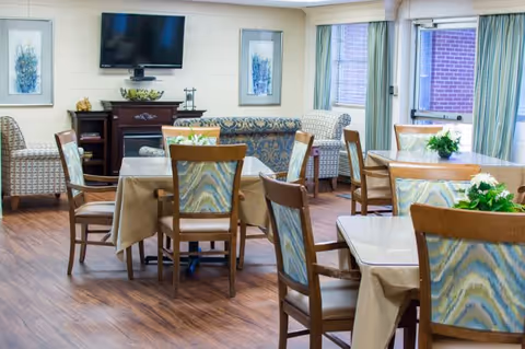 A bright and cozy common area with several tables and chairs arranged for dining or socializing. The room features wooden flooring, a wall-mounted flat-screen TV, framed artwork, and comfortable upholstered armchairs and sofas near the windows with light green curtains.