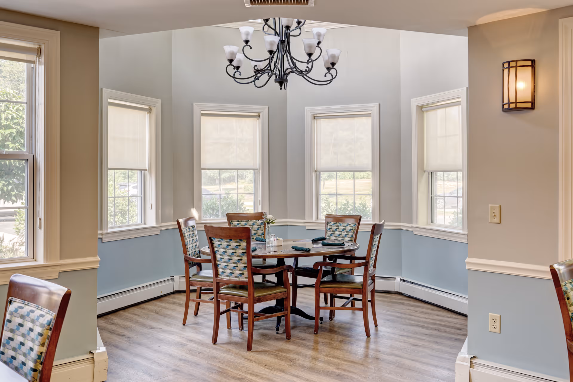 Sunlit dining area with a round wooden table surrounded by chairs, a chandelier, and three bay windows.
