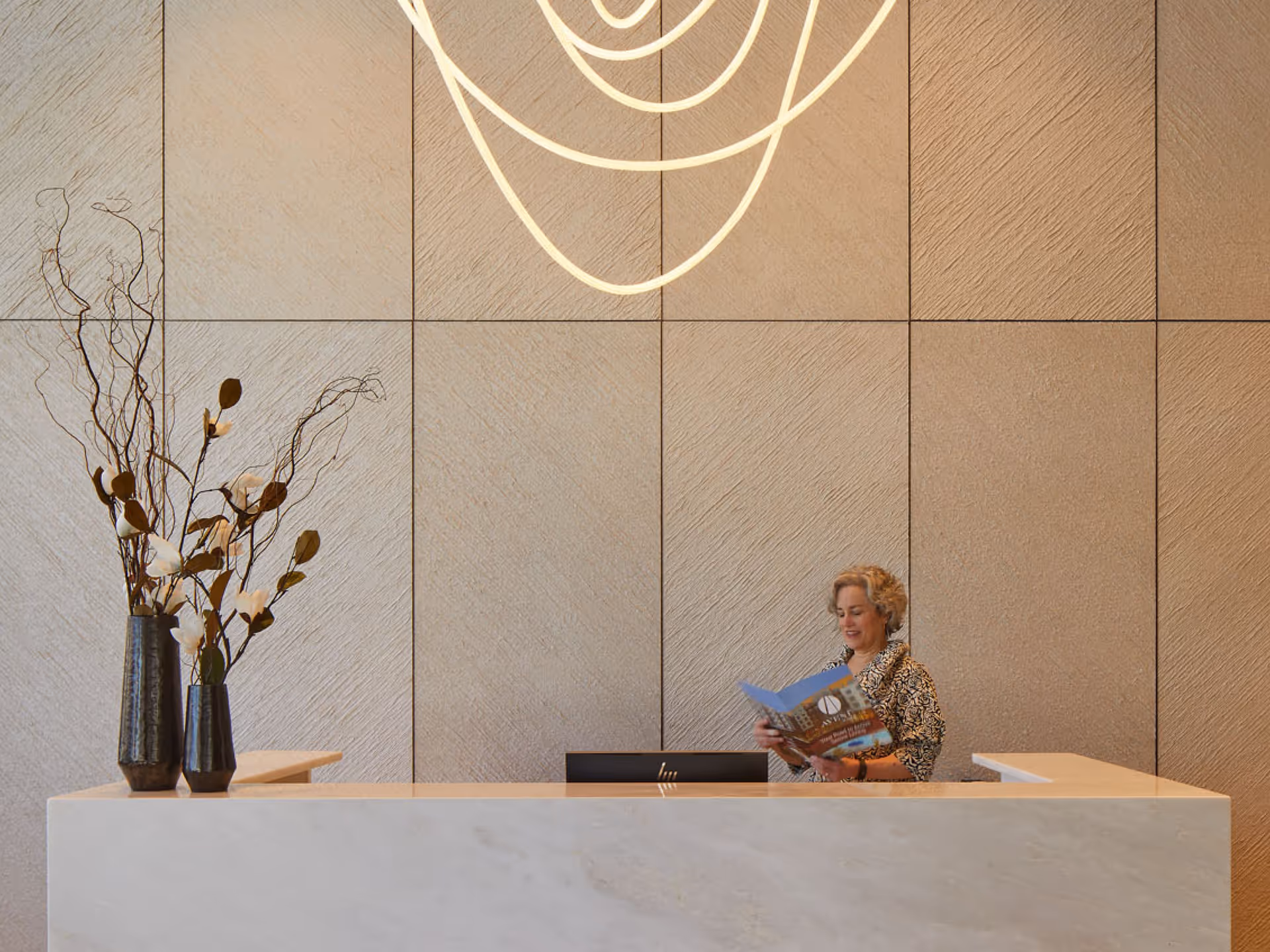 A woman standing behind a modern reception desk reading a brochure. The background features large textured beige wall panels and a contemporary light fixture above. Two tall vases with decorative branches are placed on the left side of the desk.