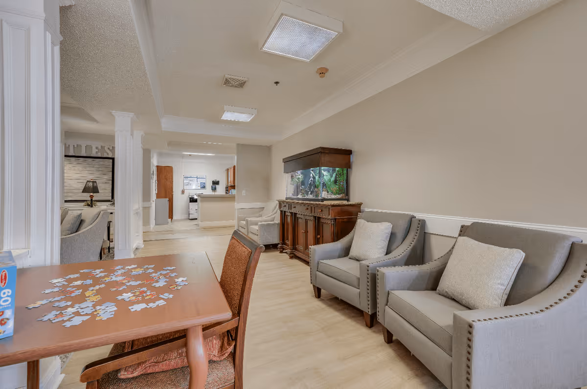 Interior view of a senior living facility lounge area with two gray armchairs with cushions, a wooden cabinet with a large fish tank on top, and a wooden table with a partially completed jigsaw puzzle and chairs around it. The background shows a kitchen area and additional seating.