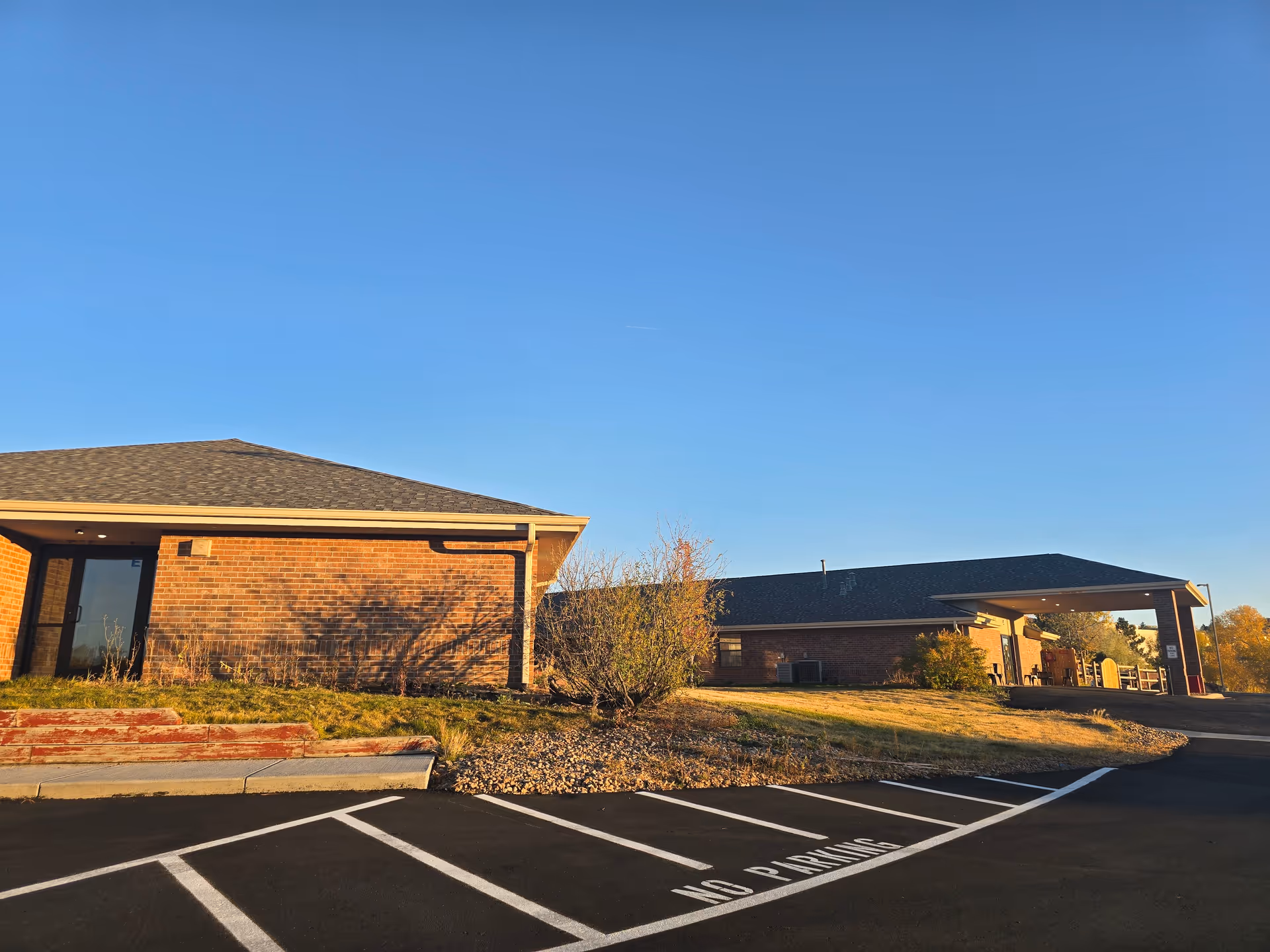 Single-story brick building exterior with a covered entrance, landscaped lawn and marked parking spaces under a clear blue sky.