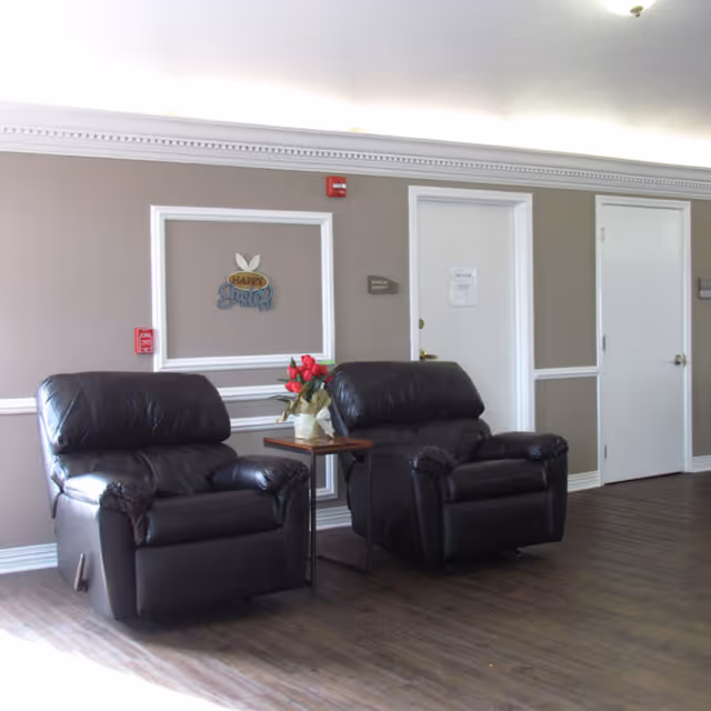 Two black leather recliner chairs with a small wooden table between them holding a vase with red flowers, set against a beige wall with white trim. There are two white doors and a decorative wall sign that says 'Happy Easter'.