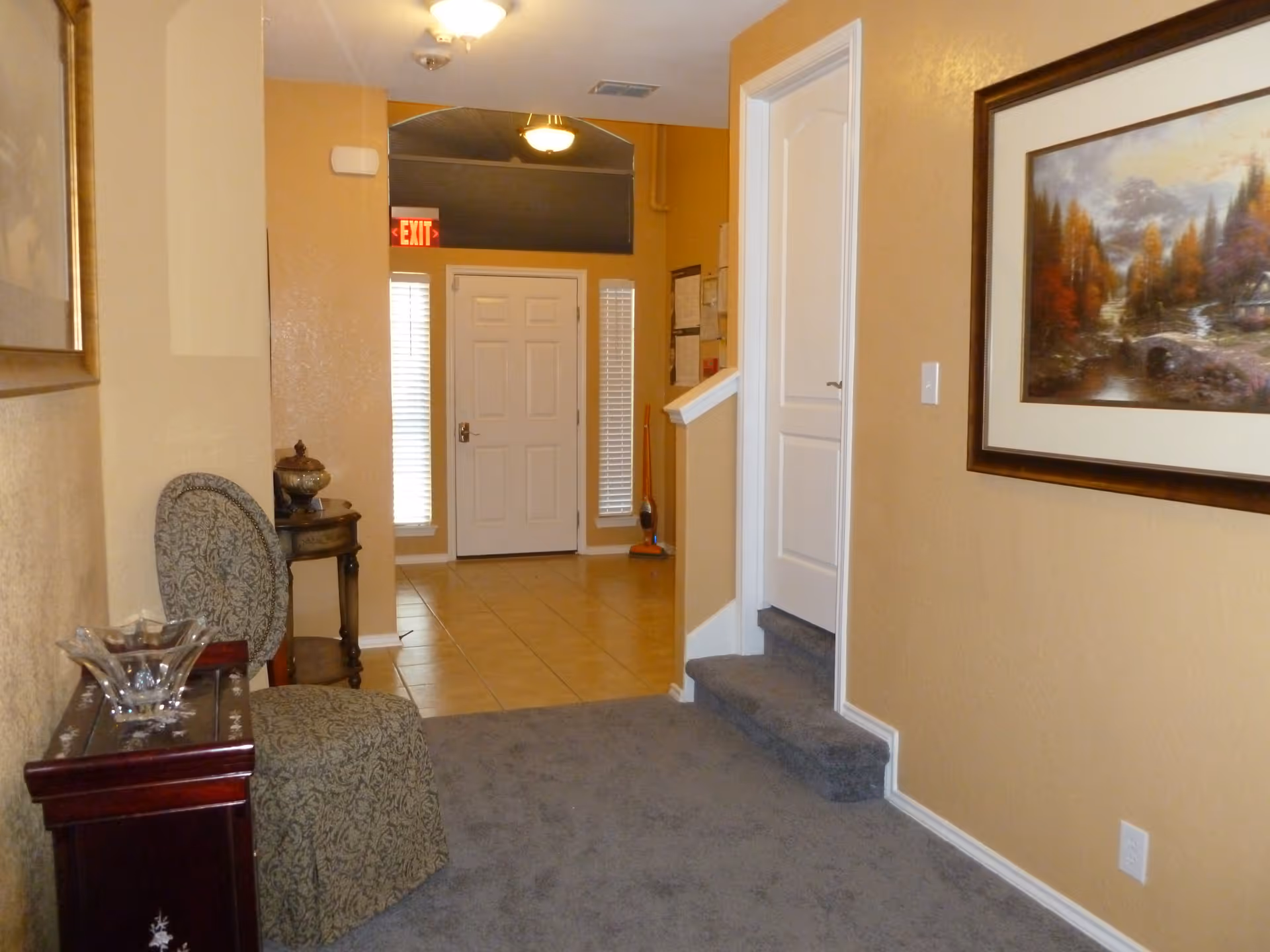 Interior hallway of a home care facility with beige walls and carpeted floor leading to a tiled entryway with a white front door. There is a patterned chair and small wooden tables with decorative items on the left side, a framed landscape painting on the right wall, and a small set of carpeted stairs leading up to a white door on the right.