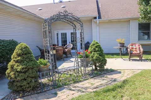 Outdoor patio area at Vintage Park at Gardner featuring a decorative metal arch with flower pots, two small evergreen shrubs, a paved walkway, and wicker chairs with cushions arranged near a building with white siding and a glass door.