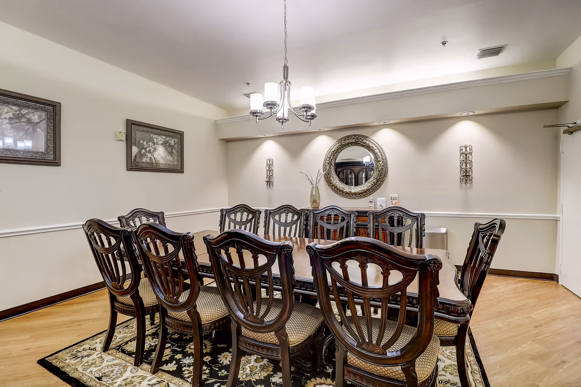 A formal dining room with a long wooden dining table surrounded by ten matching wooden chairs with cushioned seats. The room features a decorative area rug under the table, a chandelier hanging from the ceiling, a round ornate mirror on the wall, two framed pictures, and wall sconces providing accent lighting.