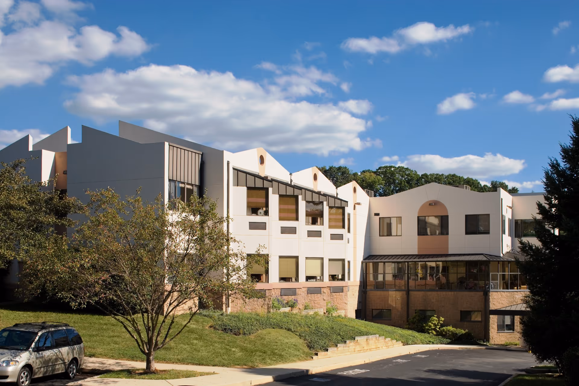 Exterior view of a multi-story building with modern architecture under a blue sky with scattered clouds. The building is surrounded by greenery including trees and bushes, with a parked car visible on the left side.