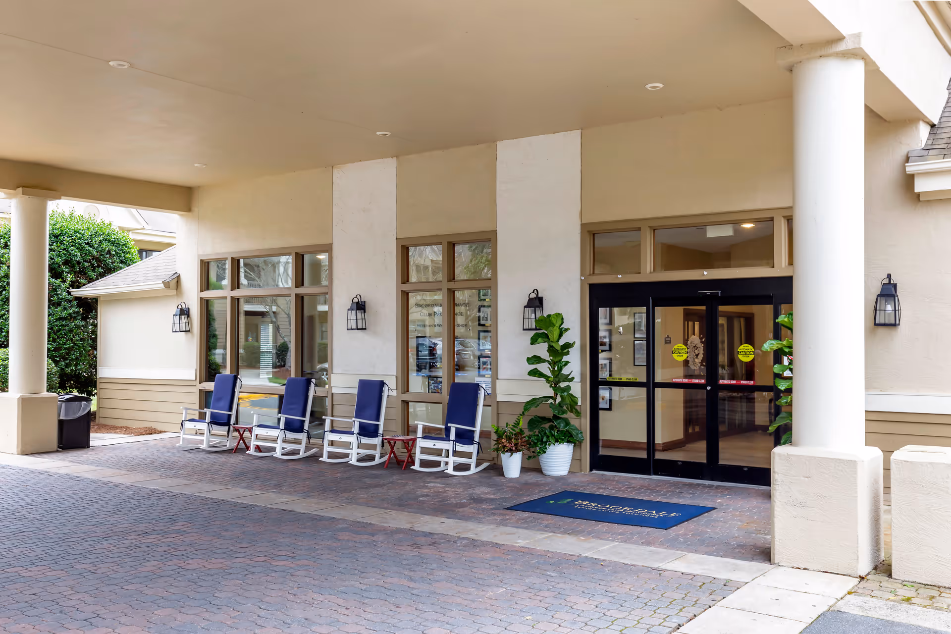 Covered entrance area of a senior living facility with four blue rocking chairs and two small red tables lined up against the wall. There are two potted plants near the glass double doors leading inside. The exterior walls are beige with large windows and black wall-mounted lantern lights.