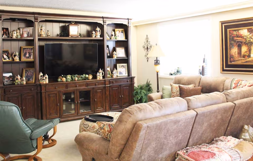 A cozy living room with a large brown sectional sofa, a green recliner chair, and a wooden entertainment center with a flat-screen TV. The shelves of the entertainment center are decorated with framed photos and ceramic pitchers. There is a floor lamp next to the sofa and a framed painting on the wall near a window letting in natural light.