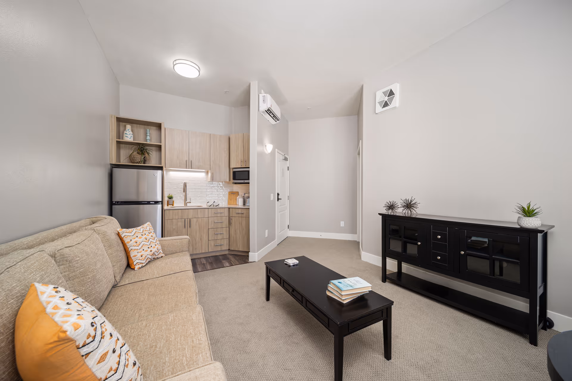 A modern living room area with a beige sofa adorned with patterned orange and white pillows. In front of the sofa is a black coffee table with a few books and a remote control on it. To the right, there is a black console table with decorative items and a small plant. The background features a compact kitchen with light wood cabinets, a stainless steel refrigerator, a microwave, and a sink. The walls are painted light gray, and the floor is carpeted except for the kitchen area which has wood flooring.