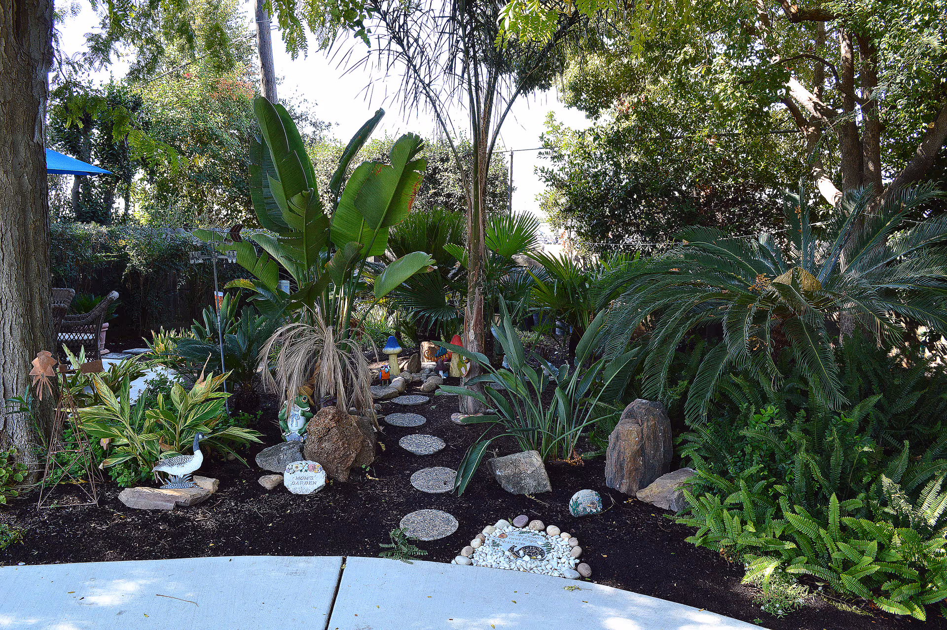 A landscaped garden path with stepping stones surrounded by tropical plants, rocks, and decorative ornaments.