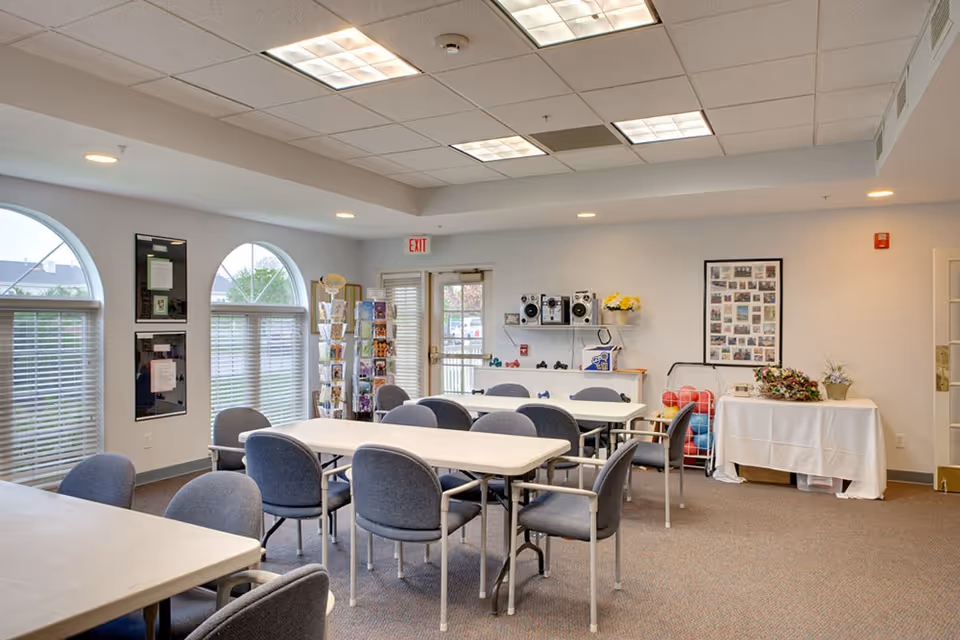 A bright communal activity room with several tables and upholstered chairs, arched windows, and a display shelf near an exit door.