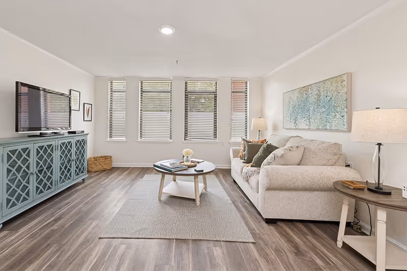 A bright and spacious living room with a light beige sofa adorned with multiple cushions, a round wooden coffee table with books and a flower vase, a blue cabinet with a flat-screen TV on top, and large windows with blinds letting in natural light. The room has wood flooring, a neutral area rug, and two table lamps providing additional lighting. A large abstract painting hangs on the wall above the sofa.