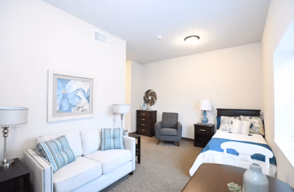 A bright and cozy assisted living room featuring a white couch with patterned blue and white pillows, two floor lamps, a framed blue floral painting on the wall, a gray armchair, a dark wooden dresser, a nightstand with a blue lamp, and a single bed with blue and white bedding near a window.