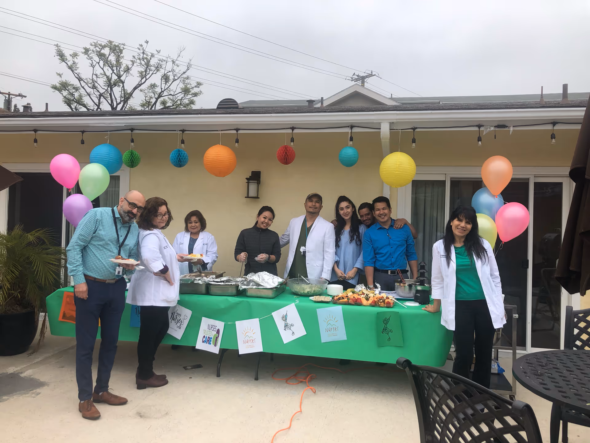 A group of eight people, some wearing white medical coats, standing behind a table covered with a green tablecloth and various food trays. The setting is outdoors with colorful paper lanterns and balloons hanging above and around the table. The background shows a building with sliding glass doors and a beige wall.