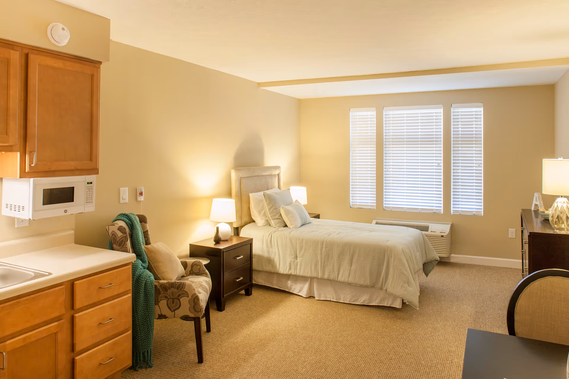 A well-lit senior living facility room featuring a single bed with pillows and a light green bedspread, two bedside tables with lamps, a patterned armchair with a green throw blanket, a kitchenette area with wooden cabinets, a microwave, and a sink, and three large windows with white blinds.
