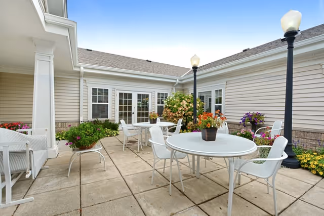 Sunny outdoor courtyard patio with round tables and white chairs, potted flowers, lampposts, and French doors on a beige-sided building.