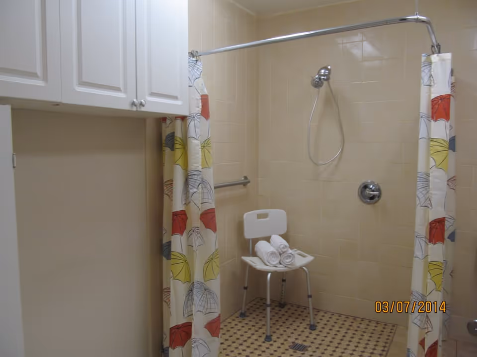 Shower area with beige tiled walls and floor, a white shower chair with three rolled white towels on it, a colorful shower curtain with umbrella patterns, and white cabinets above a beige wall.