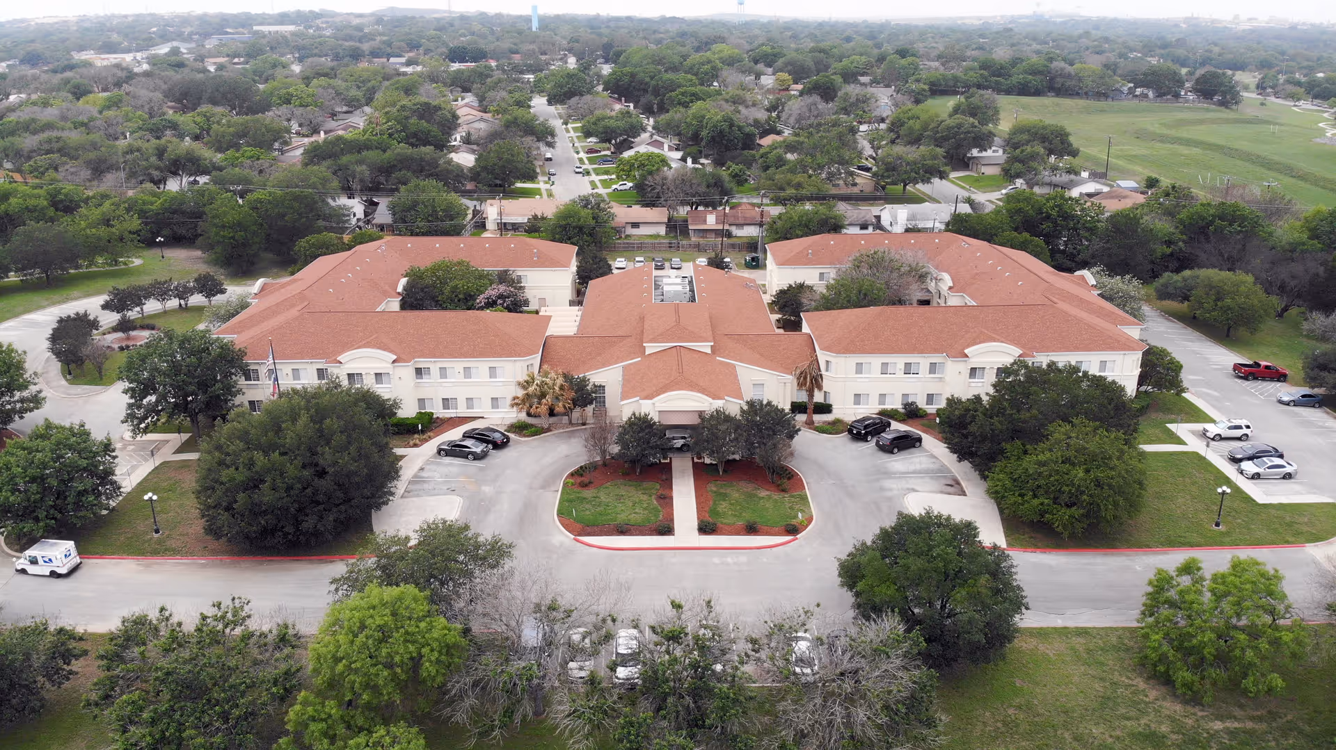 Aerial view of Esplanade Gardens senior living facility showing a large, two-story building with a red roof surrounded by trees and parking areas. The building is situated in a suburban neighborhood with many trees and houses visible in the background.