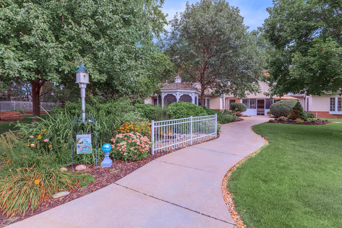 A paved walkway curves through a well-maintained garden with green grass, colorful flowers, and various trees. A white gazebo is visible in the background near a building with large windows and a brick base.