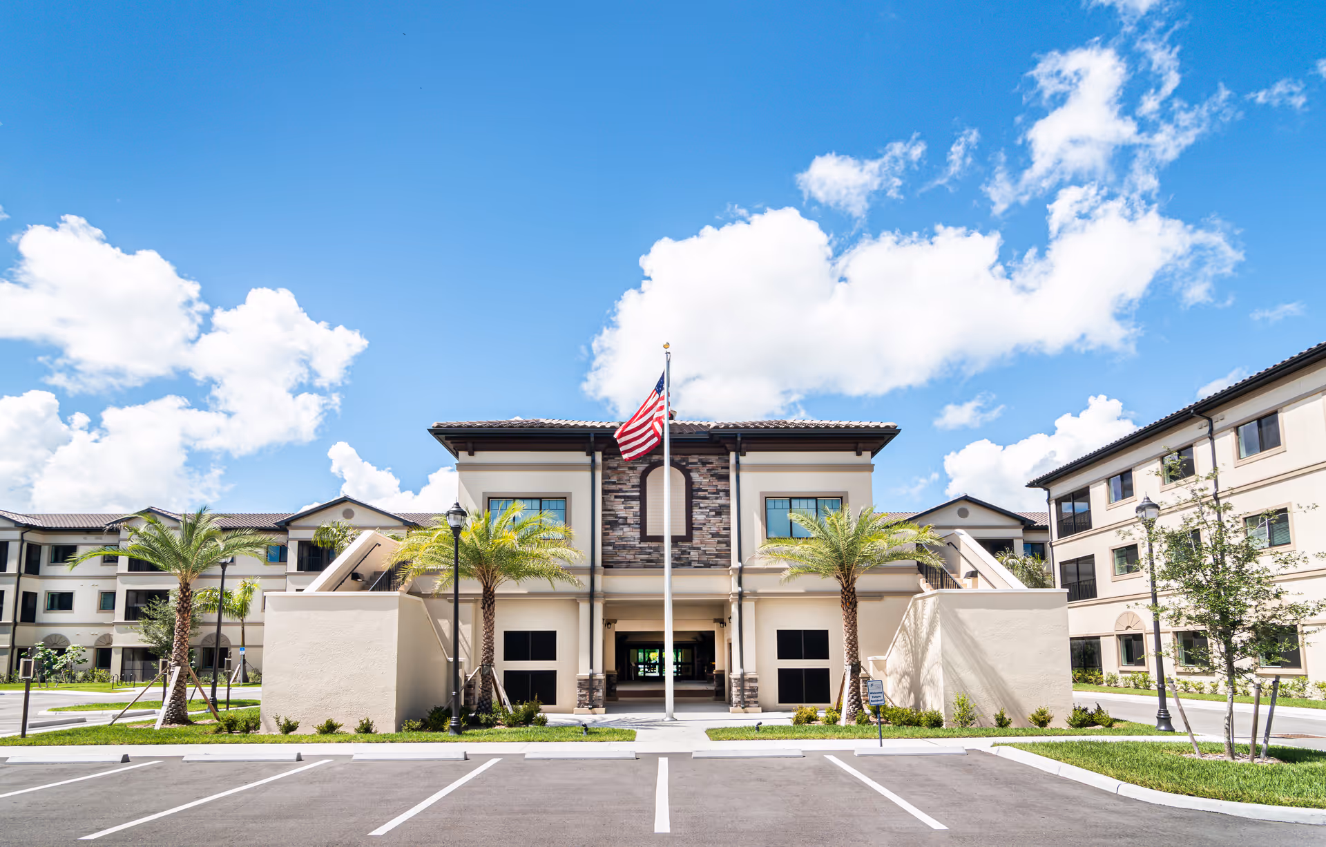 Front exterior view of a modern senior living facility building with an American flag on a flagpole in the center, palm trees on either side, and a clear blue sky with scattered clouds above.