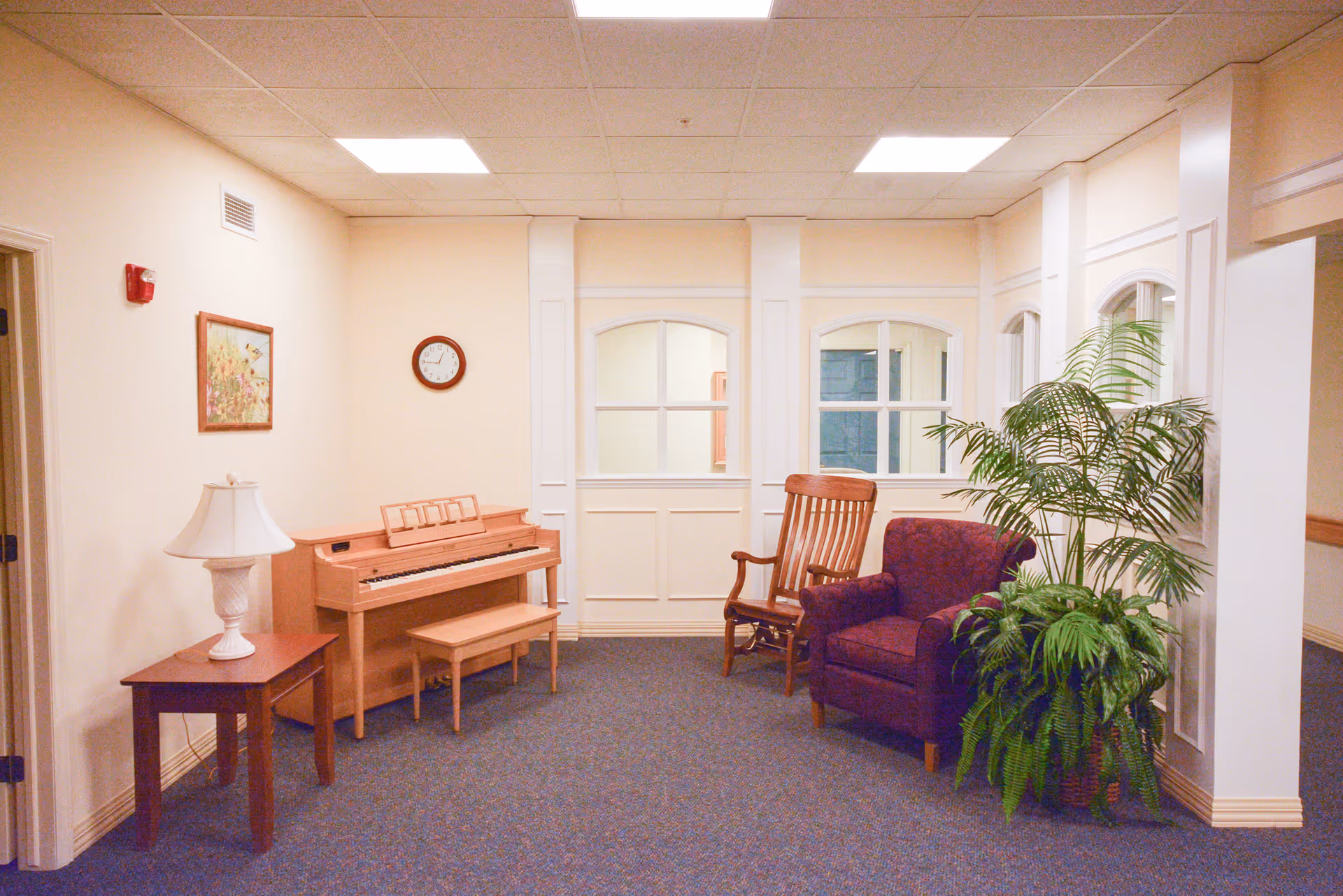 A cozy interior room with a wooden piano and matching bench against a cream-colored wall. A small wooden side table with a white lamp is next to the piano. On the wall above the piano, there is a round clock and a framed floral painting. To the right, there is a wooden rocking chair and a purple upholstered armchair next to a large green potted plant. The room has a blue carpet and white paneled walls with windows looking into another room.