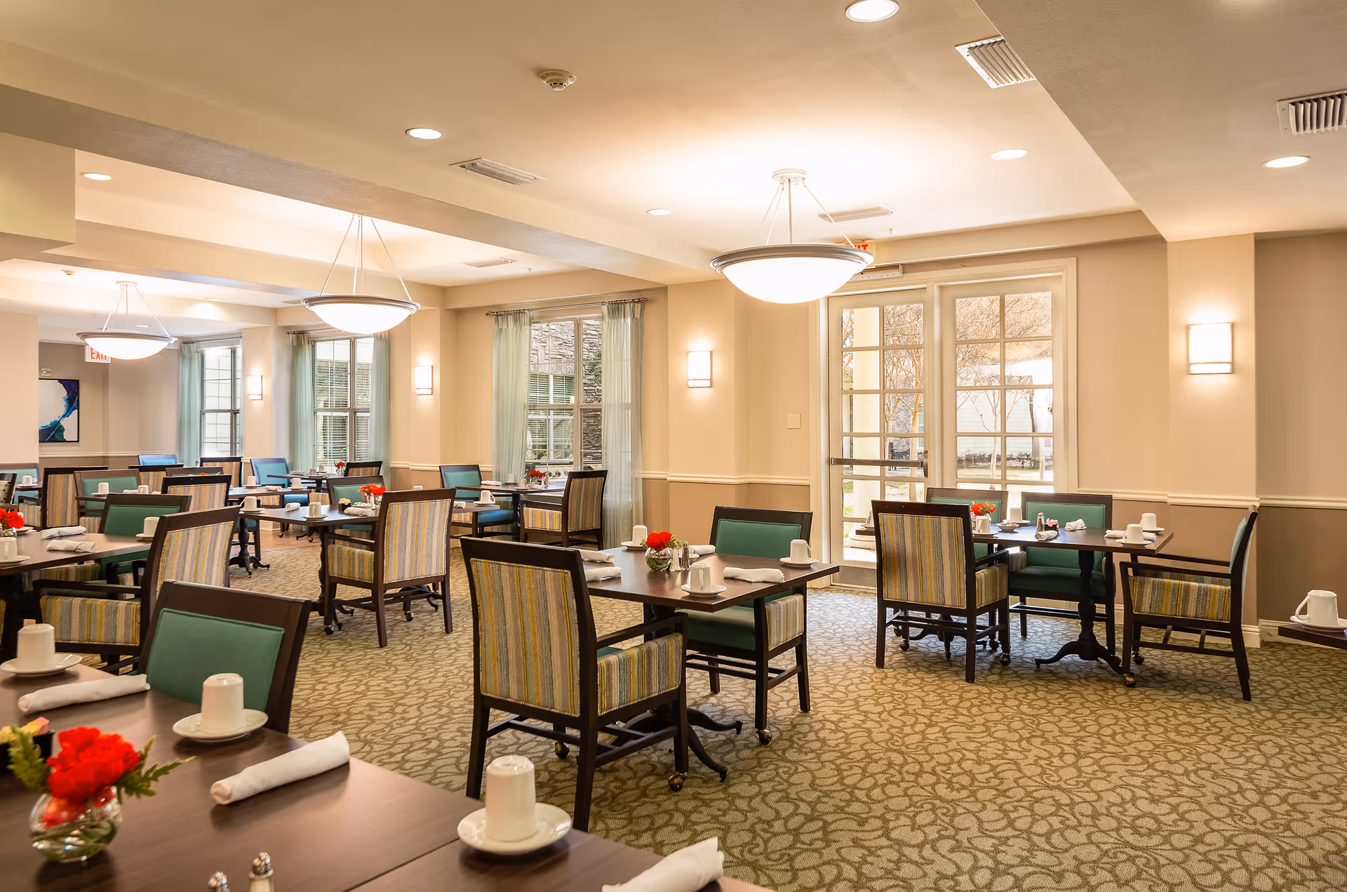 A bright and spacious dining room with multiple tables and chairs arranged neatly. Each table is set with white cups, saucers, napkins, and small vases with red flowers. Large windows with light green curtains allow natural light to fill the room. The walls are painted in a soft beige color, and modern ceiling lights provide additional illumination.