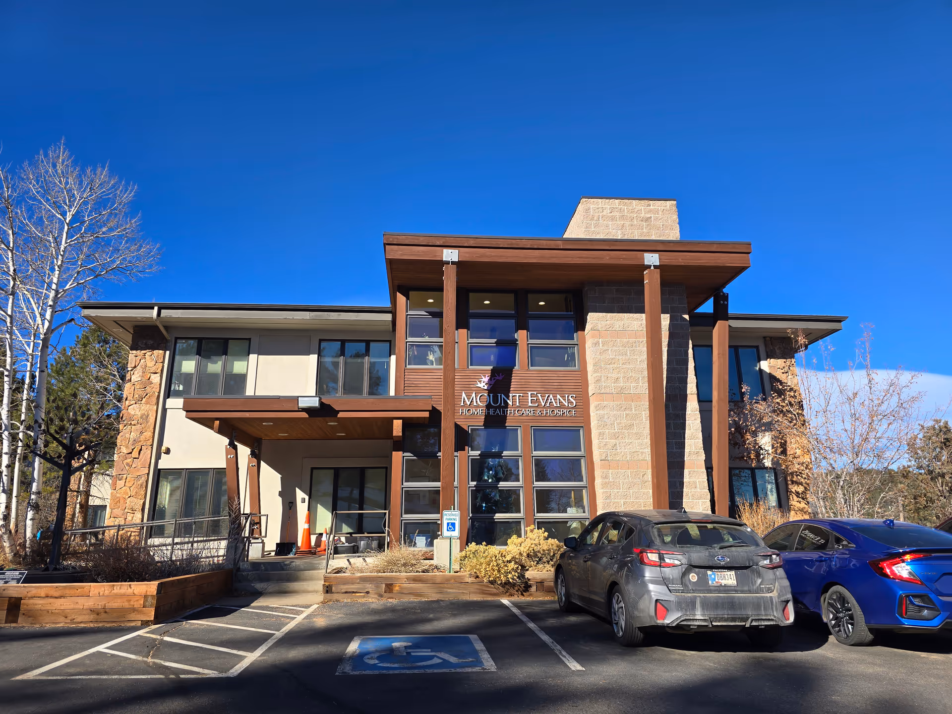 Exterior view of a two-story building with a modern design featuring stone and wood accents. There are two cars parked in front, one in a handicapped parking space. The building has large windows and a covered entrance. A sign on the building reads 'Mount Evans Home Health Care & Hospice'. Trees with no leaves are visible on the left and right sides under a clear blue sky.