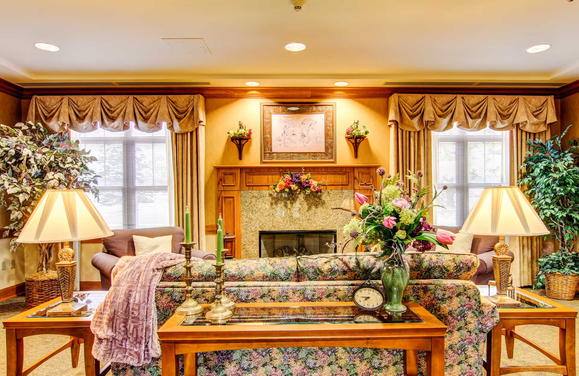 A cozy living room with a floral patterned sofa facing a fireplace with a wooden mantel. The mantel is decorated with flowers and a framed picture above it. Two armchairs with pillows are positioned near windows with beige curtains and valances. Two table lamps with beige shades sit on wooden side tables on either side of the sofa. There are also two green plants in wicker baskets on each side of the room, and a glass-top coffee table with a vase of flowers and decorative items in front of the sofa.