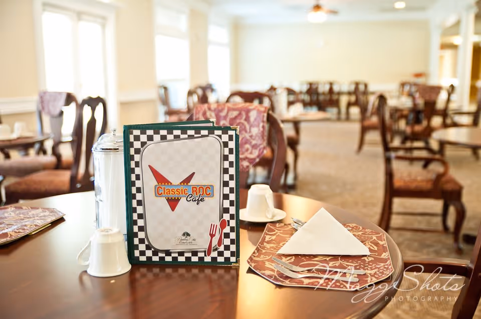 A dining table set with a menu titled Classic ROC Cafe, a folded napkin on a decorative placemat, a cup, and a sugar container in a well-lit dining room with multiple tables and chairs in the background.