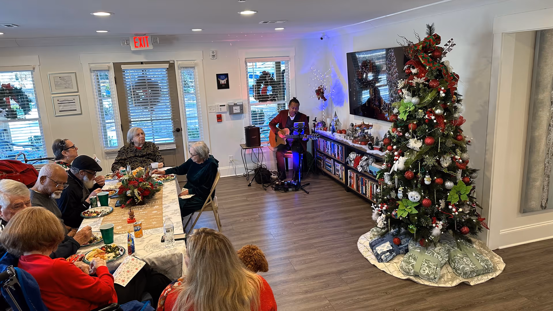 Residents seated at a long table eating in a decorated common room while a musician plays guitar next to a large Christmas tree.