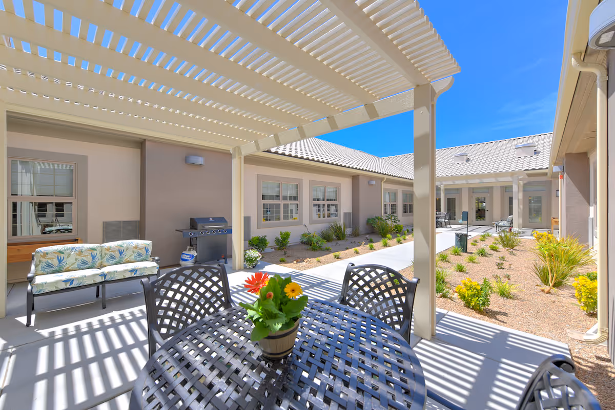 Shaded outdoor courtyard patio with a metal table and chairs, potted flowers, a bench, grill, and surrounding single-story memory care building.