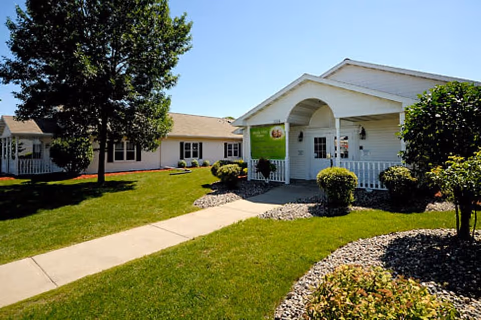 Exterior view of Marla Vista Assisted Living and Memory Care facility showing a white building with a covered entrance, surrounded by green lawn, bushes, and trees under a clear blue sky.