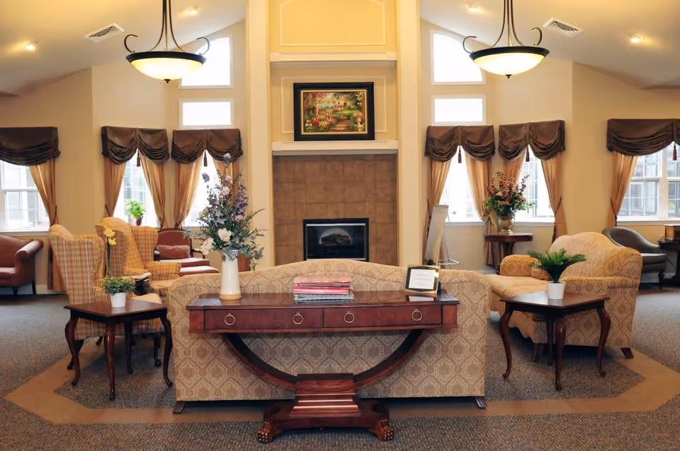 A cozy and elegant living room area in Encore Memory Care at Bolingbrook featuring a central fireplace with a framed painting above it, surrounded by multiple windows with brown curtains. The room is furnished with patterned sofas, armchairs, wooden side tables with potted plants and floral arrangements, and a wooden console table with books and a small framed sign.