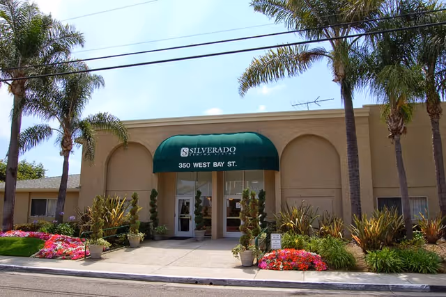 Front exterior view of Silverado Newport Mesa Memory Care Community building with a green awning displaying the name and address 350 West Bay St. The entrance is flanked by potted plants and surrounded by palm trees and colorful flower beds.