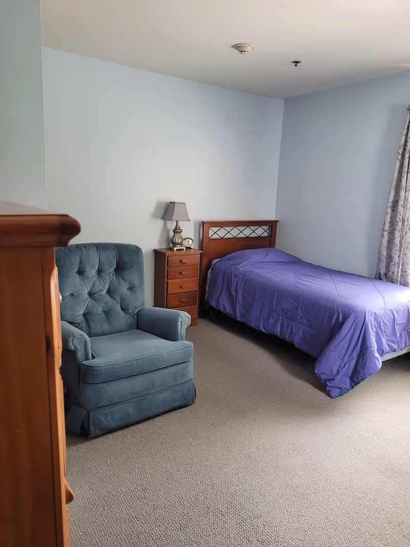 A simple bedroom with a single bed covered in a purple bedspread, a wooden nightstand with a lamp and a clock, a blue upholstered armchair, and light blue walls with a window covered by patterned curtains.