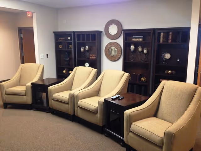 A seating area with four beige upholstered armchairs arranged in a row, separated by dark wooden side tables. Behind the chairs are two dark wooden shelving units with decorative items such as clocks, vases, and small sculptures. Two round mirrors with textured frames are mounted on the wall between the shelving units. The room has beige carpet and neutral-colored walls.